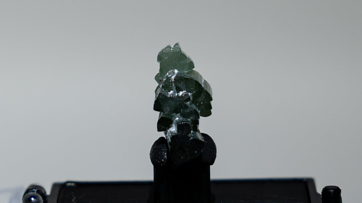 A small dark rectangular display case holding a green carved figurine, shown on a white pedestal with a clear acrylic cover partially open in the foreground against a light gray studio background.