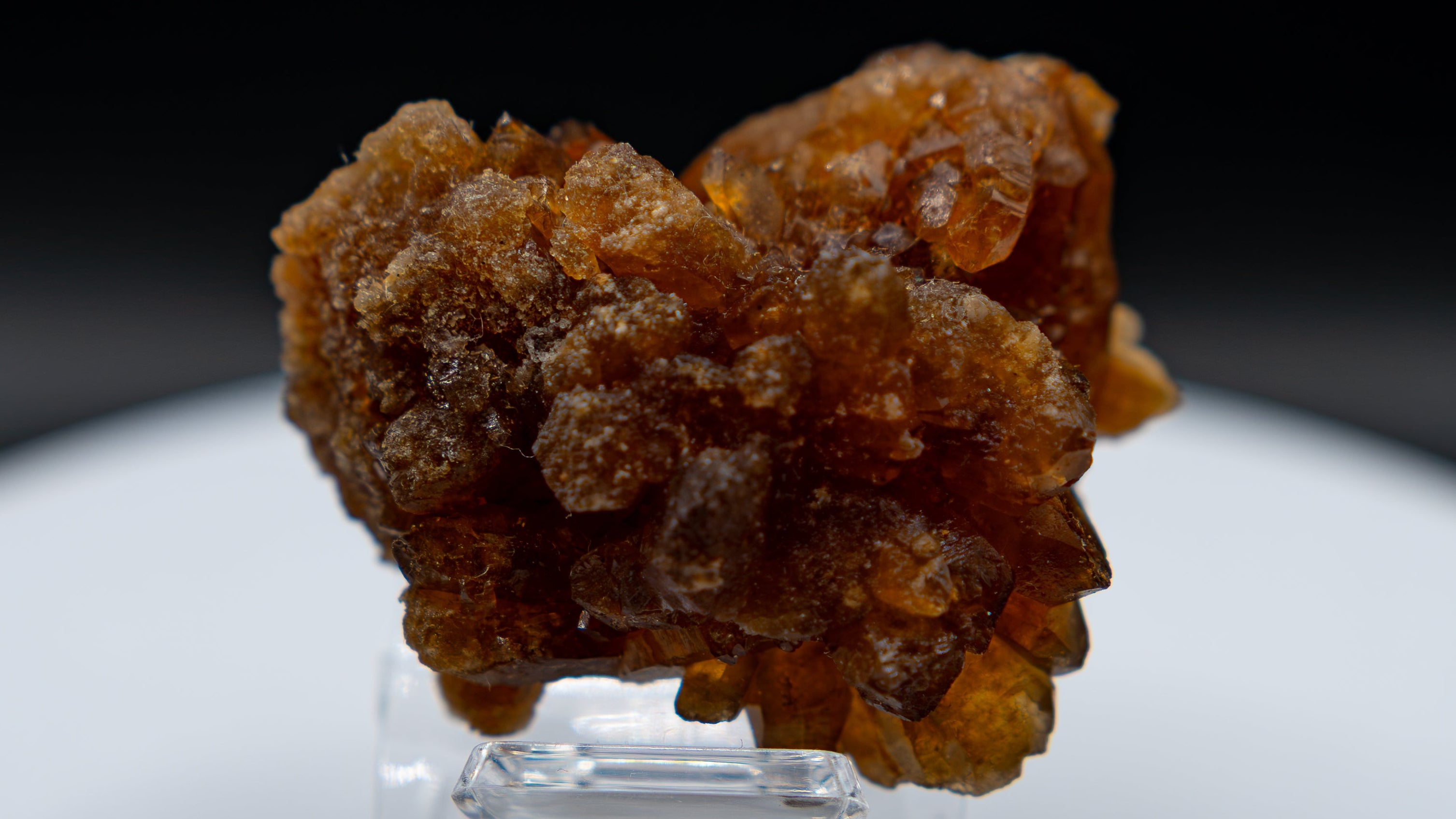 A rough, textured brown crystal cluster displayed on a small clear acrylic stand, set against a dark black background and a white circular pedestal.