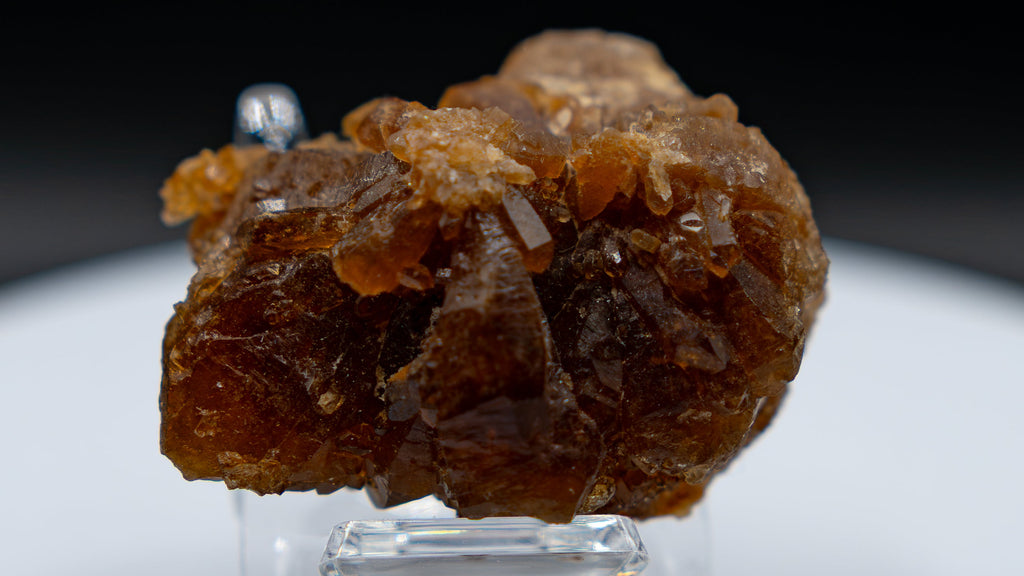 A small rough brown crystal cluster displayed on a clear stand atop a round white pedestal against a dark background.