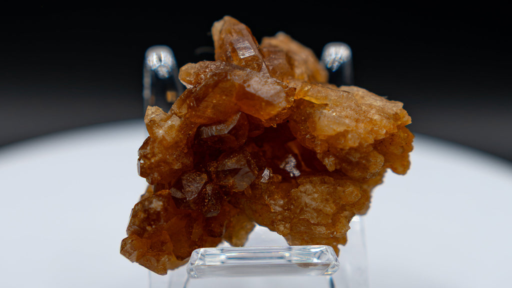 A small cluster of amber-brown translucent crystals displayed on a clear stand atop a round white pedestal against a black background.