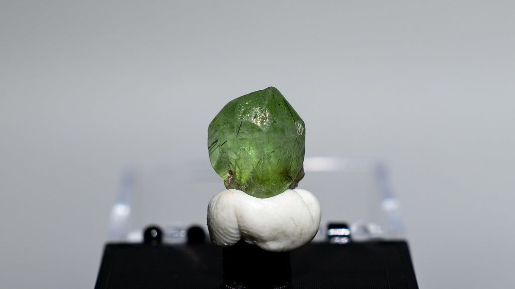 A small green gemstone or crystal mounted on a white sculptural base and displayed on a black rectangular stand, set against a light gray studio background.
