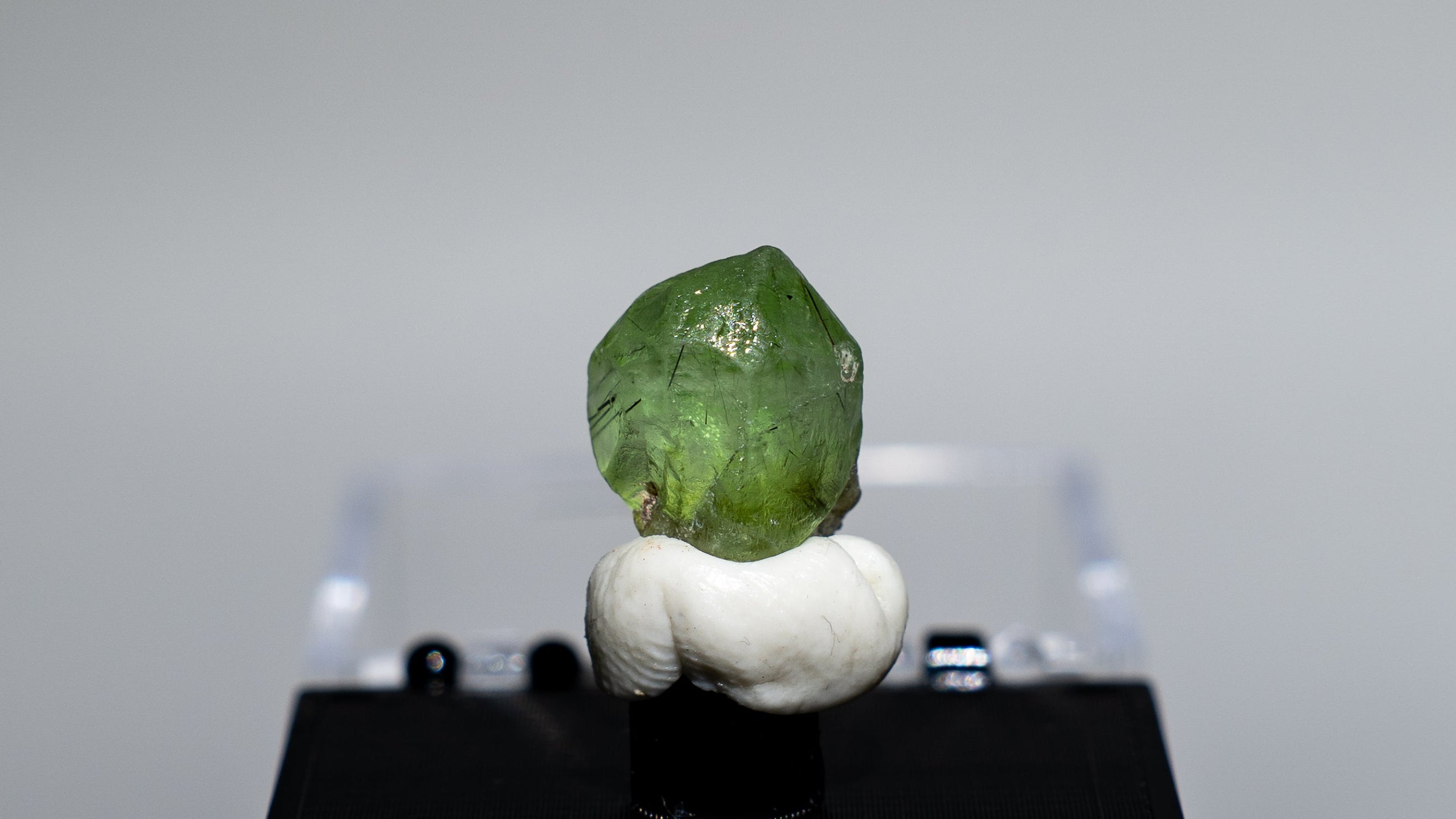 A small green gemstone or crystal mounted on a white sculptural base and displayed on a black rectangular stand, set against a light gray studio background.