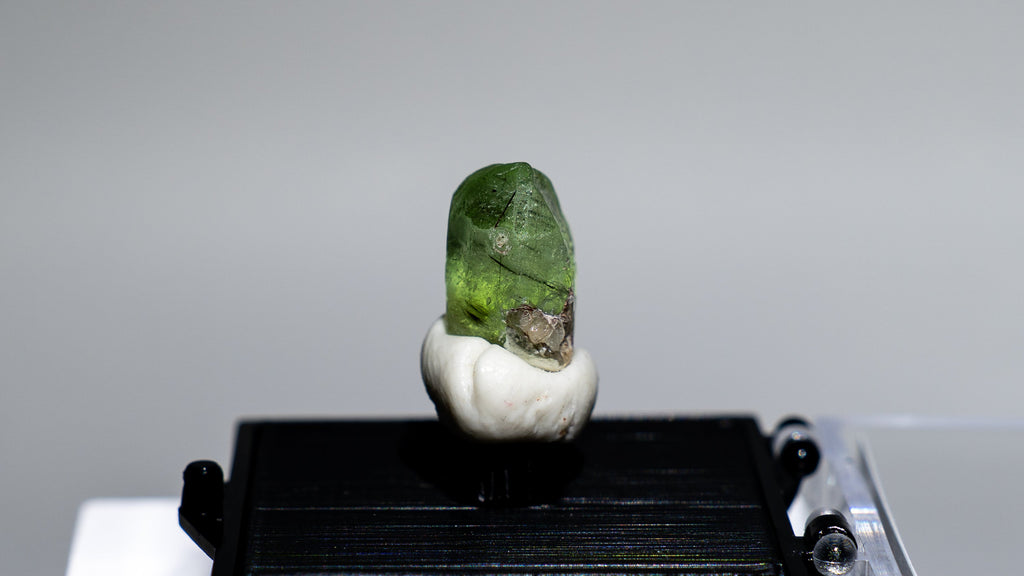 A small green gemstone or crystal mounted on a white base, displayed on a black rectangular stand inside a clear acrylic case against a light gray background.