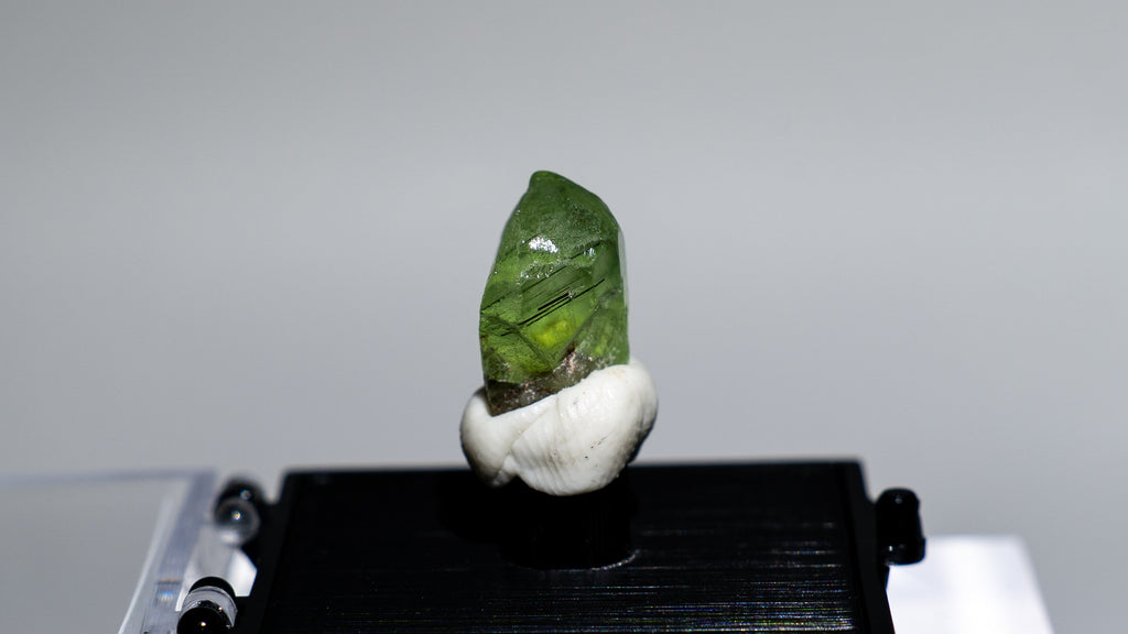 A small green polished stone or crystal mounted on a white shell-like holder, displayed on a black rectangular stand atop a clear acrylic block against a light gray background.