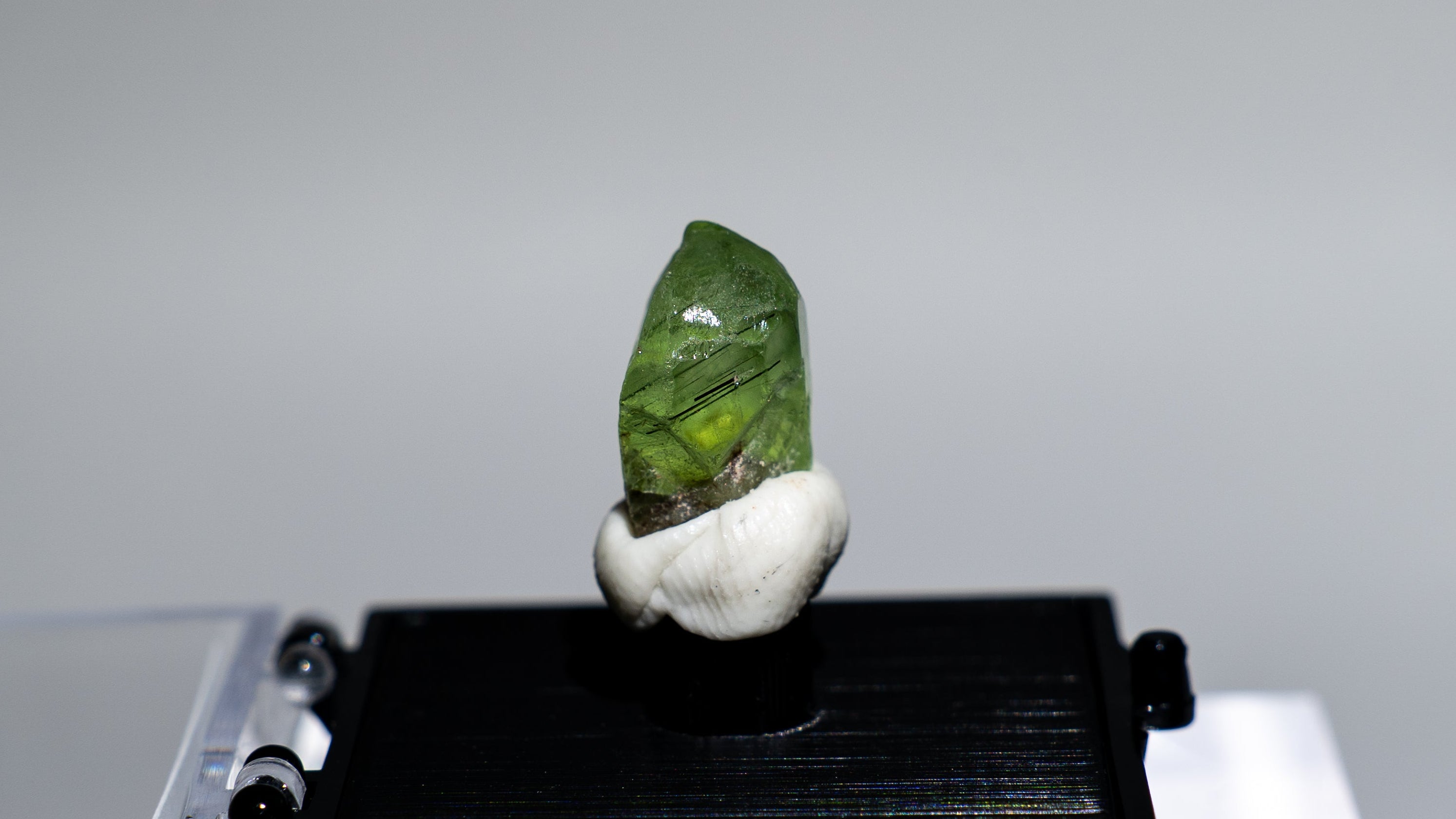 A small green polished stone or crystal mounted on a white shell-like holder, displayed on a black rectangular stand atop a clear acrylic block against a light gray background.