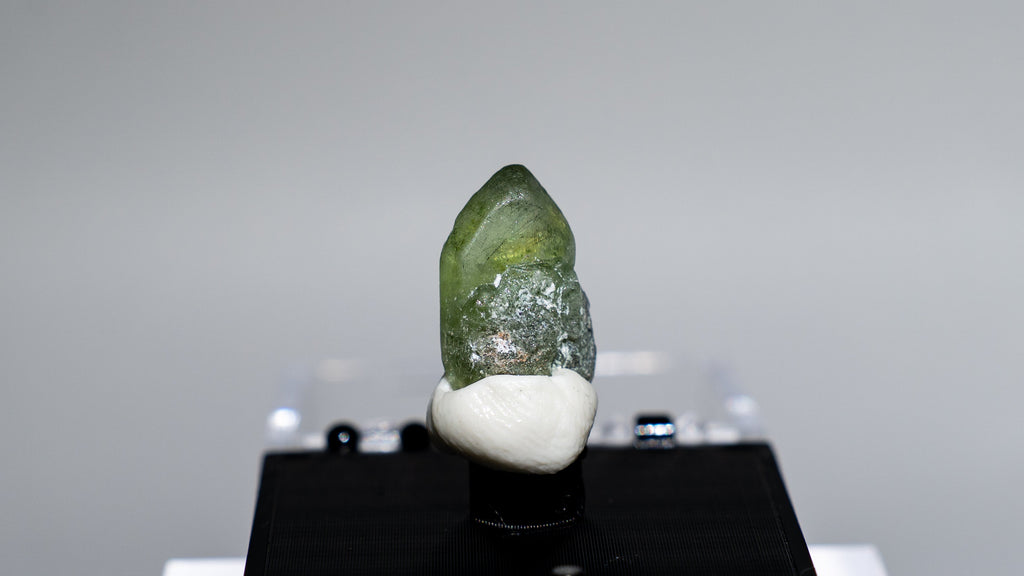 A small green and white crystal specimen mounted on a black display block, photographed against a soft gray studio background.