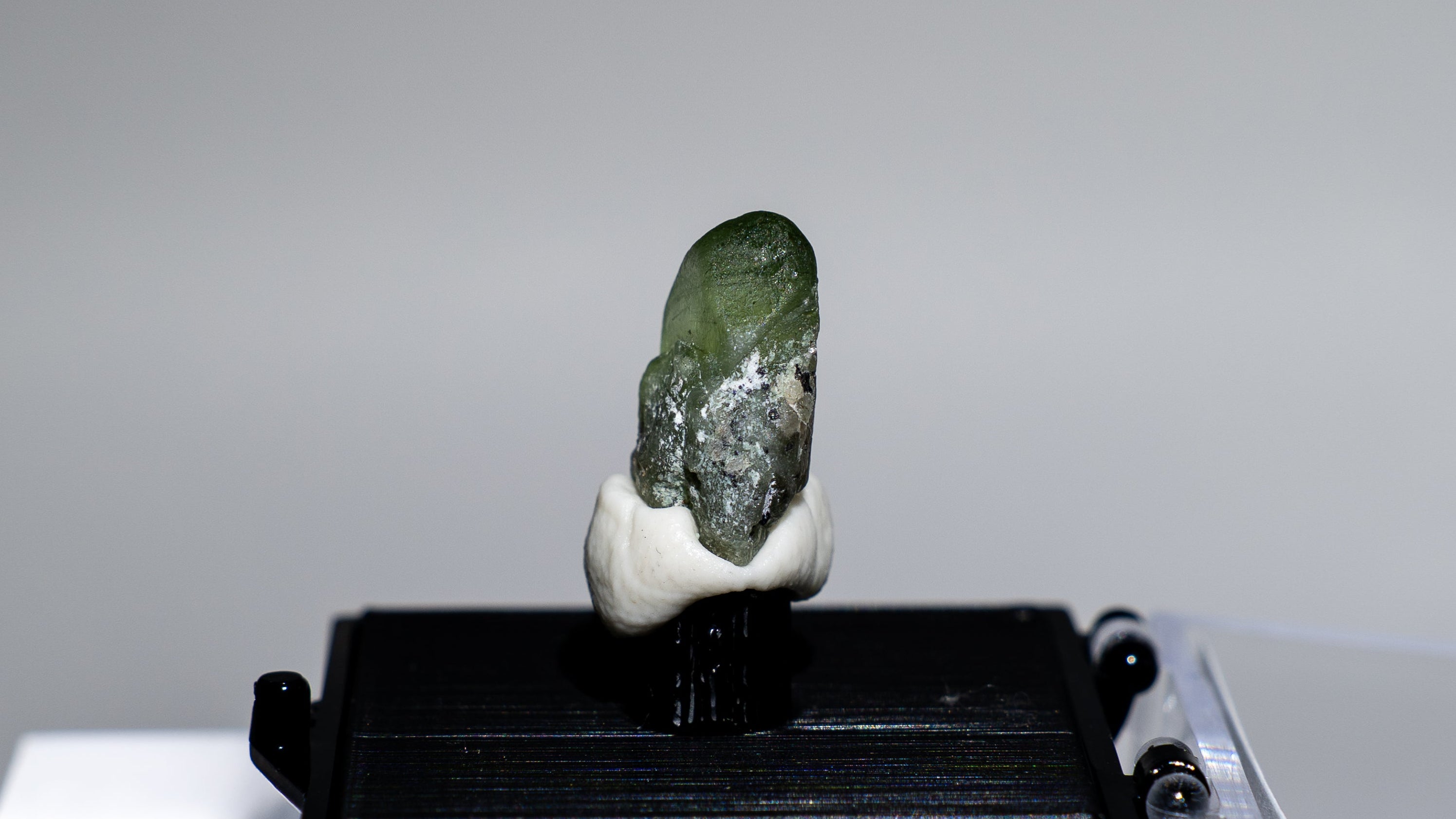 A small carved green stone figurine mounted on a black display stand, with a clear acrylic case beside it, photographed against a plain light gray background.