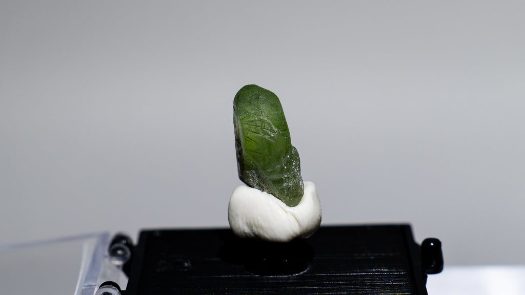 A small green crystal or mineral specimen mounted on a white support above a black rectangular base, displayed on a clear acrylic stand against a light gray background.