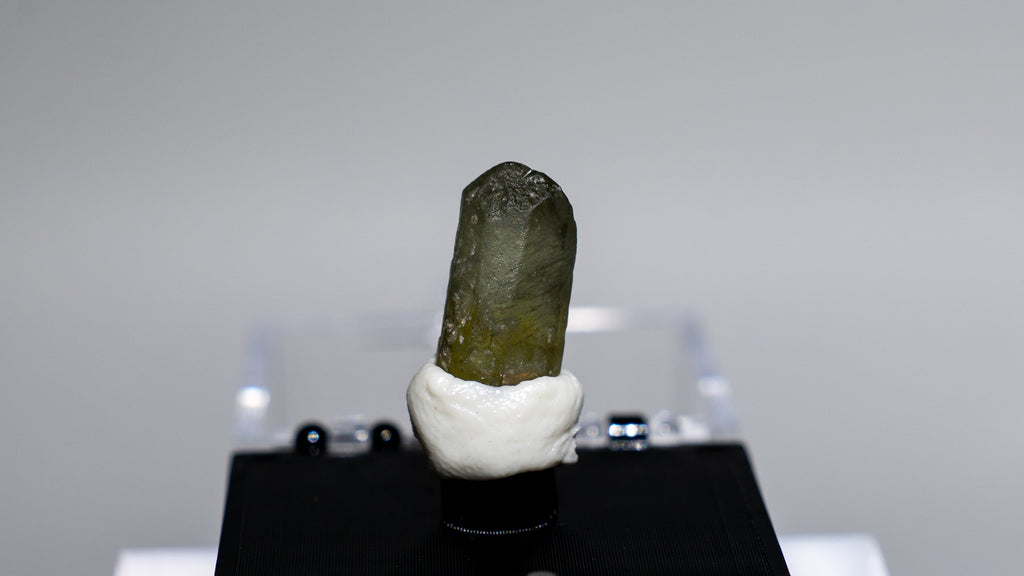 A small natural mineral or crystal specimen with a dark olive-green top and white base, mounted on a black display block on a clear acrylic stand against a light gray background.