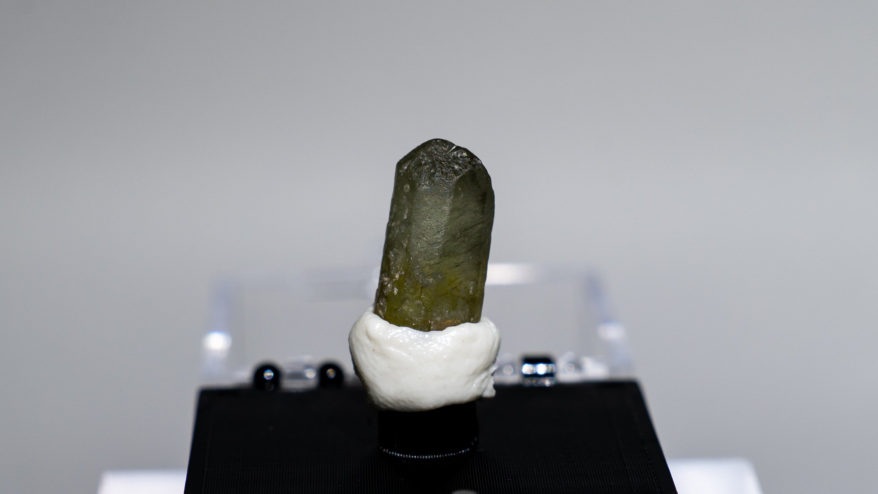 A small natural mineral or crystal specimen with a dark olive-green top and white base, mounted on a black display block on a clear acrylic stand against a light gray background.