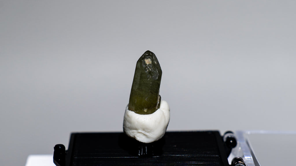 A small green crystal or mineral specimen mounted on a white stand, placed on a black display block next to a clear acrylic box, photographed against a plain light gray background.