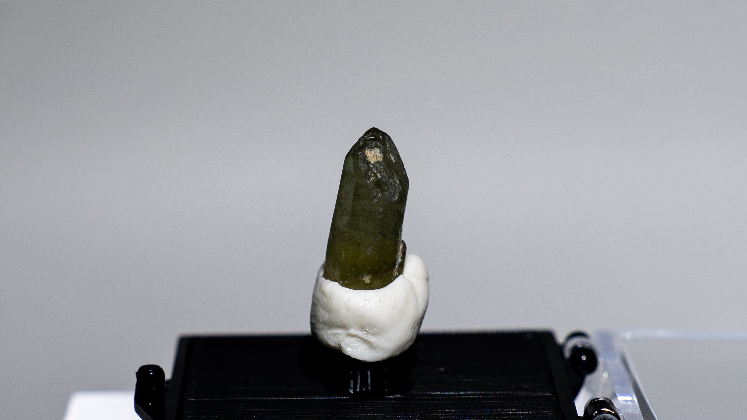 A small green crystal or mineral specimen mounted on a white stand, placed on a black display block next to a clear acrylic box, photographed against a plain light gray background.