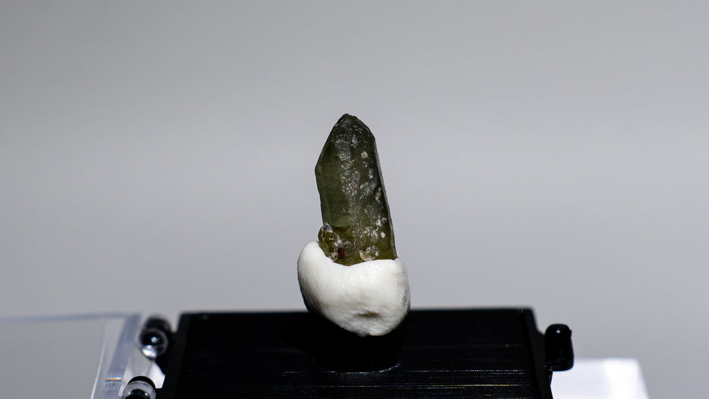 A small mineral specimen with a dark greenish crystal on a white mount, displayed on a black stand over a clear acrylic riser against a light gray background.