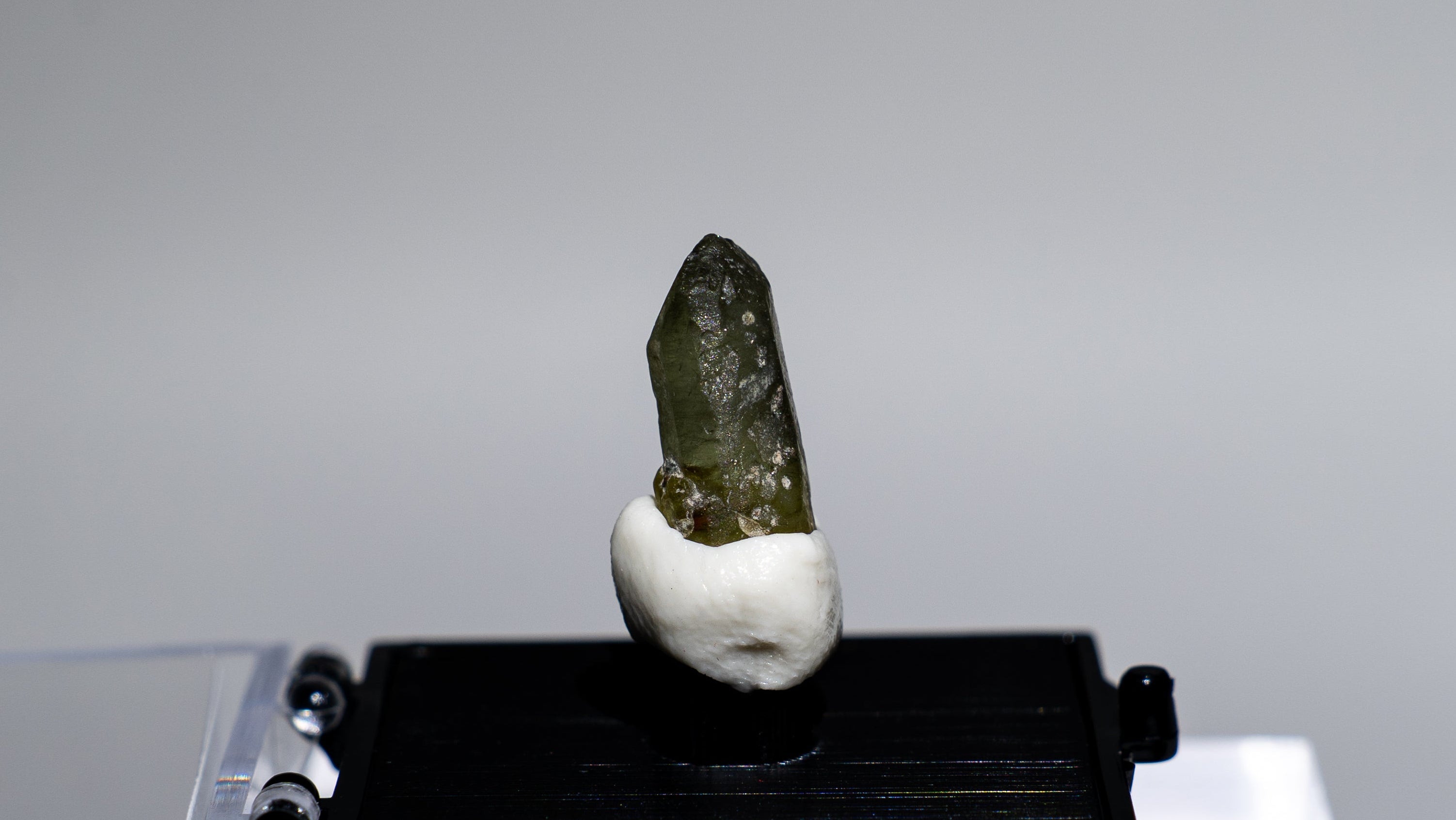 A small mineral specimen with a dark greenish crystal on a white mount, displayed on a black stand over a clear acrylic riser against a light gray background.