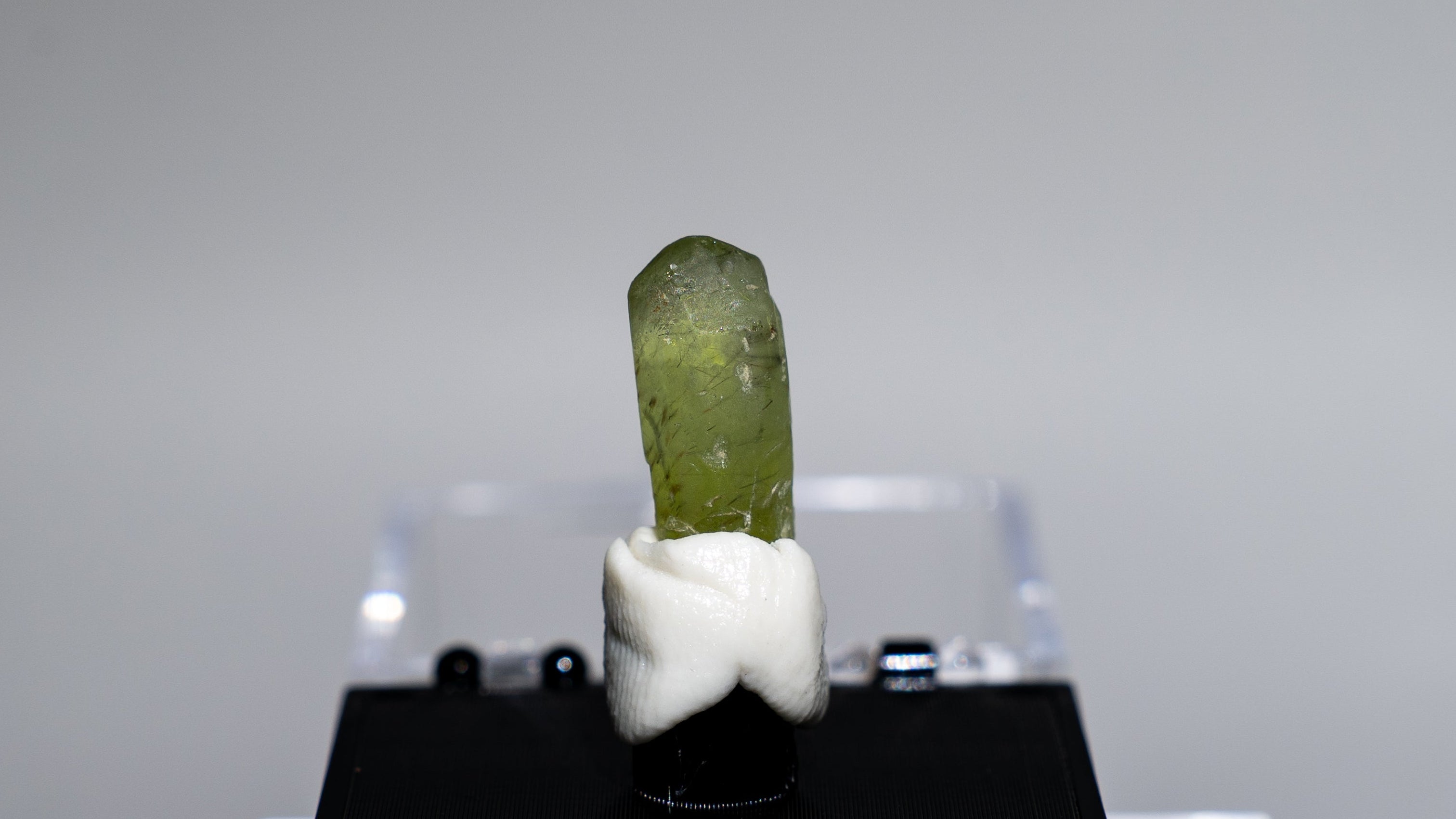 A small green crystal or mineral specimen mounted on a white block and displayed on a black stand, photographed against a plain light gray studio background.