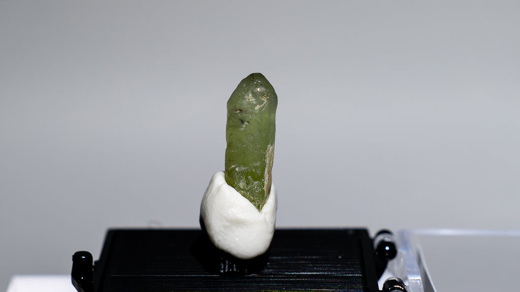 A small green crystal or mineral specimen mounted on a white base and displayed on a black rectangular stand, with a clear acrylic case beside it against a light gray background.