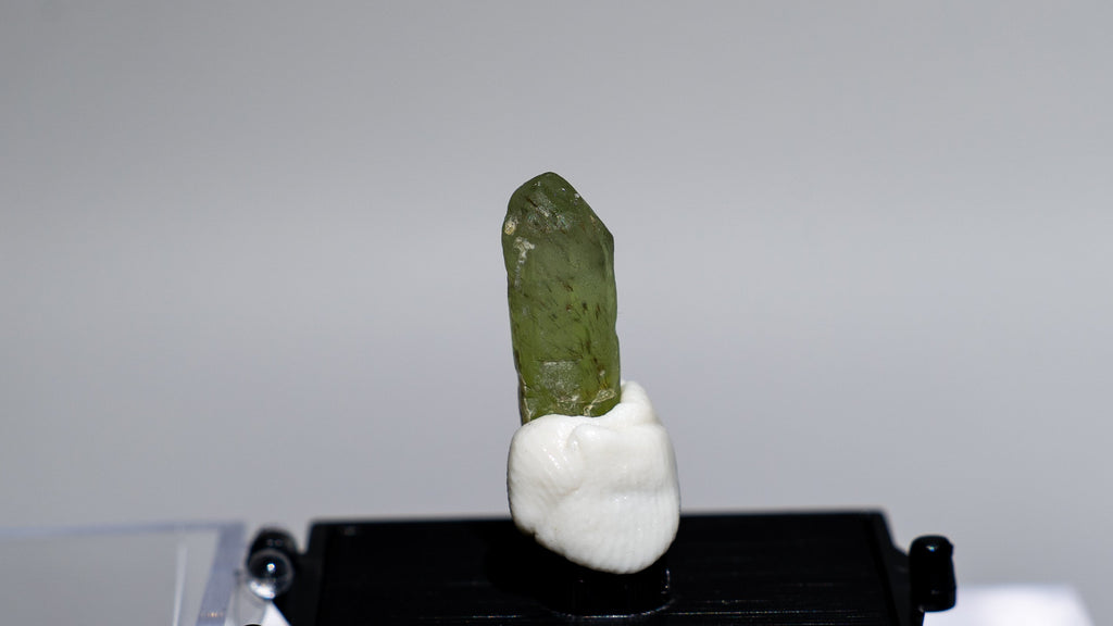 A small green crystal or mineral specimen mounted on a white base and displayed on a black rectangular stand, set against a light gray studio background with a clear acrylic display block.