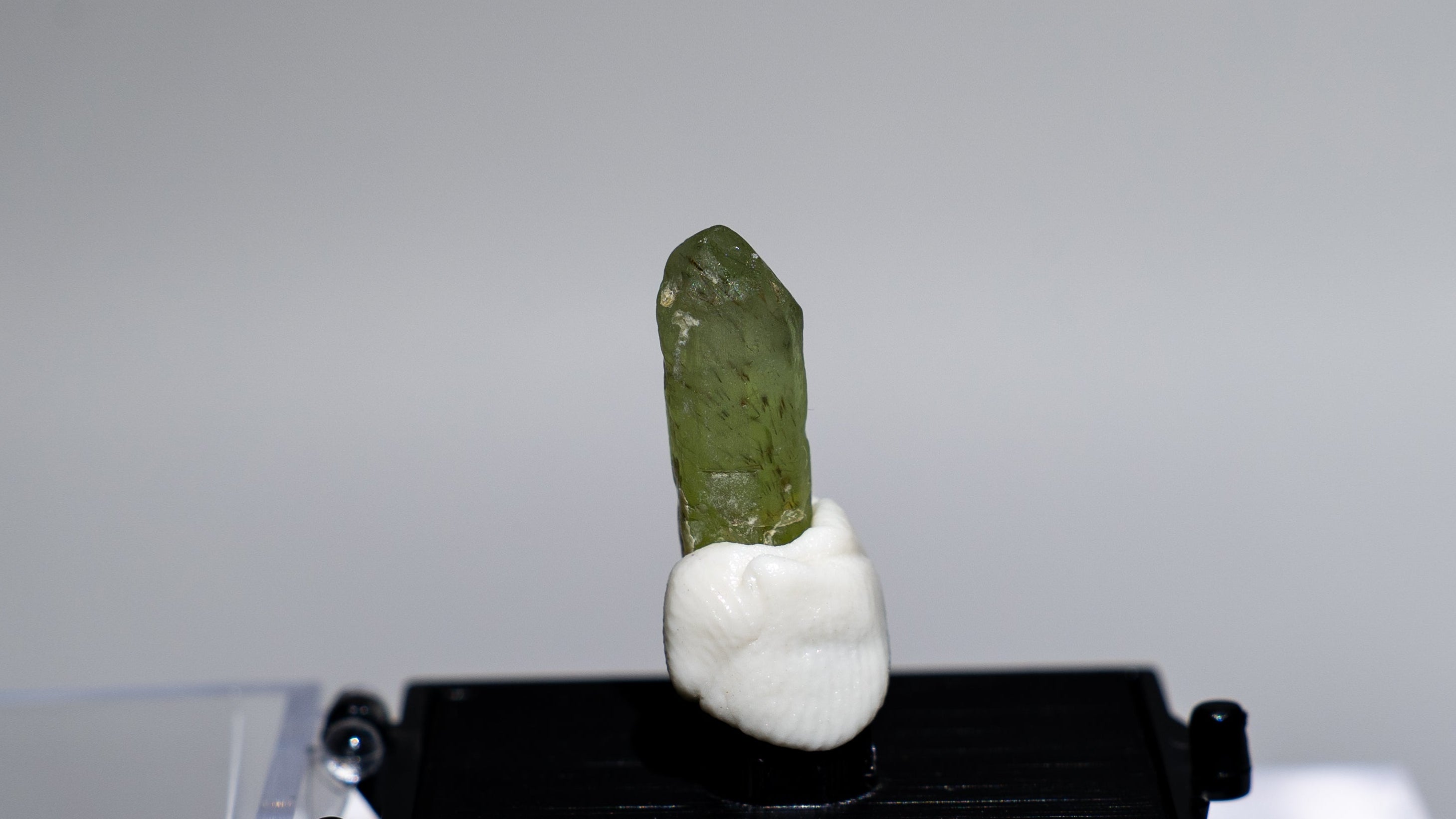 A small green crystal or mineral specimen mounted on a white base and displayed on a black rectangular stand, set against a light gray studio background with a clear acrylic display block.