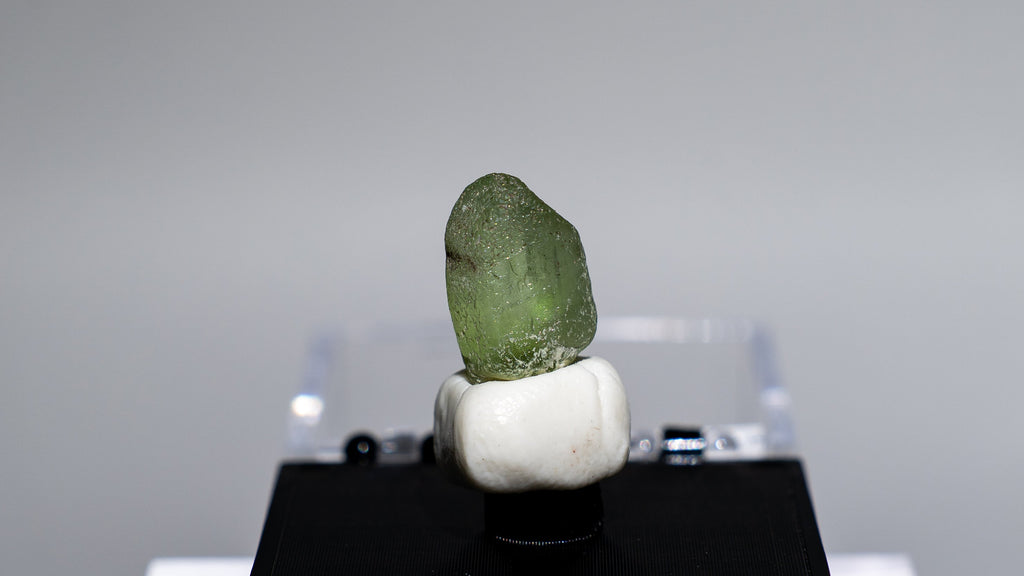 A small green stone or crystal mounted on a white round base and displayed on a black rectangular stand, set against a light gray studio background.
