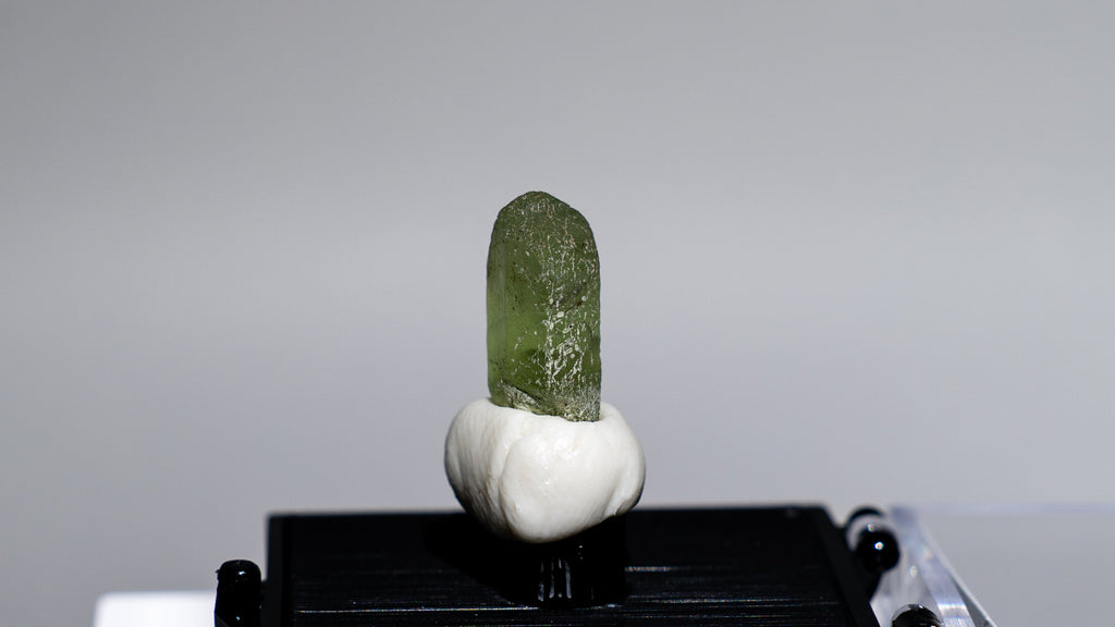 A small specimen or crystal mount displayed on a black rectangular base, with a pale green translucent top and a white bulb-like section, set against a light gray background with a clear acrylic stand visible.