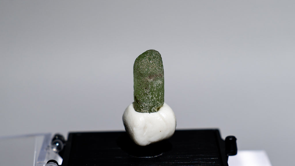 A small green stone or crystal mounted on a white base and displayed on a black rectangular platform, set against a light gray background with a clear acrylic stand.