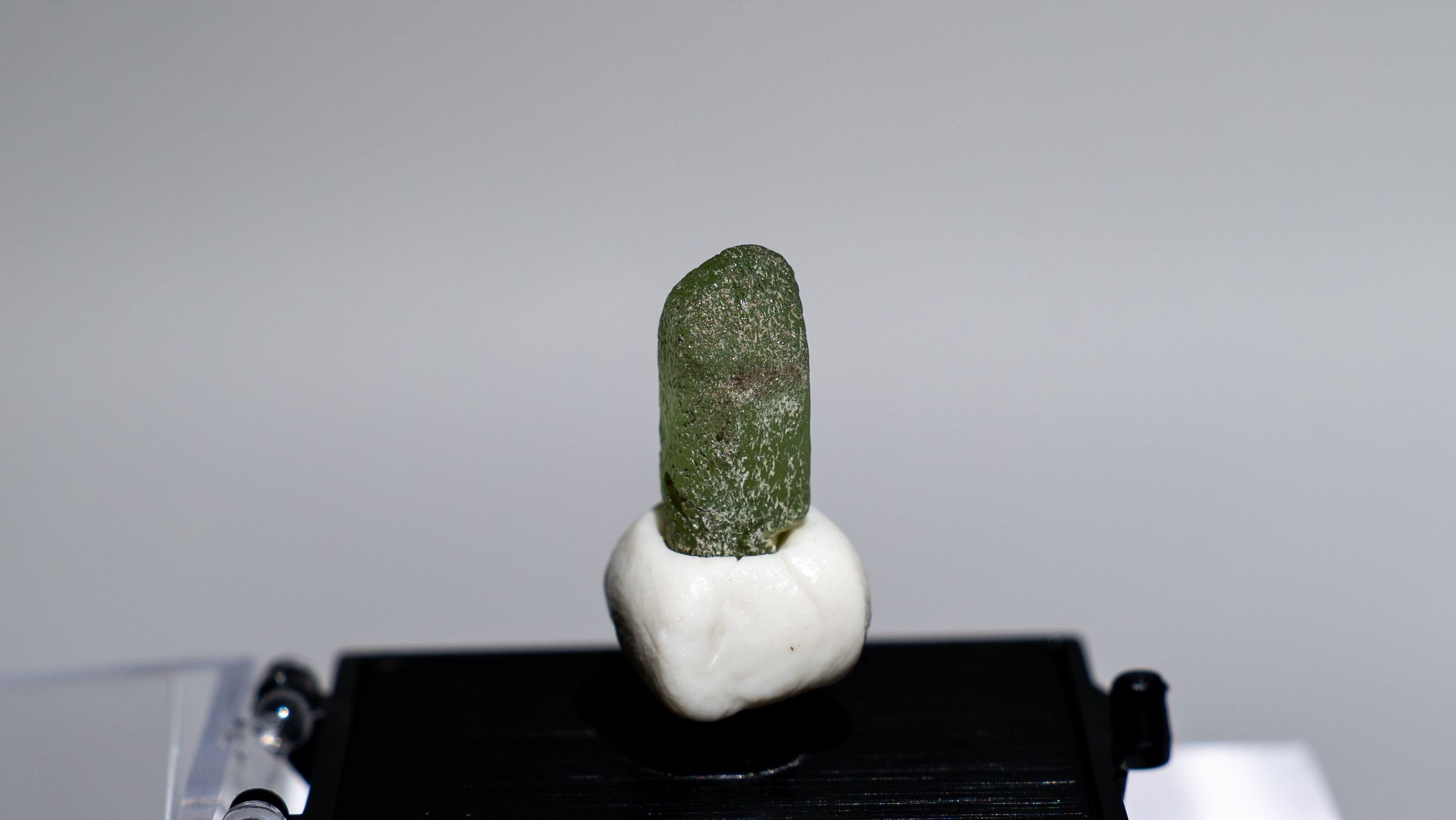 A small green stone or crystal mounted on a white base and displayed on a black rectangular platform, set against a light gray background with a clear acrylic stand.